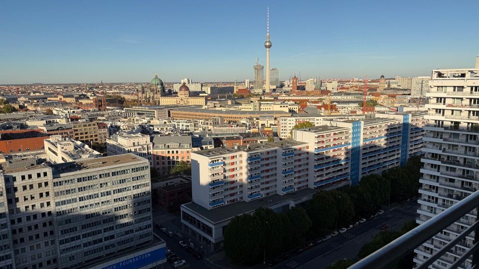 Blick über Berlin mit mehreren Wohngebäuden und Bürohäusern im Vordergrund. Im Hintergrund sind der Berliner Fernsehturm, der Berliner Dom und weitere historische Gebäude und Wahrzeichen zu sehen, unter klarem Himmel.