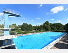 Ein Außenbereich eines Schwimmbads mit einem großen rechteckigen Becken, umgeben von Gehwegen und Grünflächen. Links steht ein blauer Sprungturm mit verschiedenen Plattformen, im Hintergrund sind Bäume und ein sonniger Himmel zu sehen.
