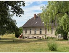 Zweistöckiges Wohngebäude mit hellem Putz und dunklem Satteldach, umgeben von einer grünen Wiese und hohen Bäumen, darunter eine hängende Weide. Im Hintergrund ist ein weiter, parkartiger Landschaftsbereich erkennbar.