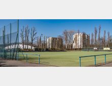Ein Sportplatz mit Kunstrasen, umgeben von blauem Geländer und hohen Zaunstrukturen. Im Hintergrund stehen mehrstöckige Wohngebäude, kahle Bäume und eine weiße, kuppelförmige Sporthalle unter einem klaren blauen Himmel.