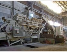 Industriehalle mit einer großen Maschine zur Materialverarbeitung, bestehend aus Förderbändern, Trichtern und einer Steuerkabine. Die Umgebung ist geprägt von Metallstrukturen, Rohren und einer Betonwand, mit einfallendem Licht von oben.