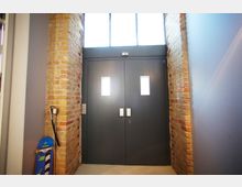 Dunkelgraue Doppeltür mit kleinen rechteckigen Fenstern, eingerahmt von unverputzten Backsteinwänden, darüber ein schmaler Fensterbereich. Im Vordergrund lehnt ein blau-orangefarbenes Skateboard an der Wand.