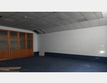 Ein leerer Raum mit einer weißen Ziegelwand, blauem Teppichboden und einer abgehängten Decke mit mehreren Lichtpaneelen. Auf der linken Seite befindet sich eine Holzwand mit mehreren Glaseinsätzen.