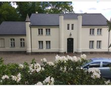 Weiß gestrichenes Gebäude mit symmetrischer Fassade, großen weißen Sprossenfenstern und zwei runden Säulen, die den Eingang mit einer dunkelgrünen Tür flankieren. Im Vordergrund sind blühende weiße Büsche und ein geparkter silberner Kleinwagen sichtbar, umgeben von einer Kiesfläche und Bäumen im Hintergrund.