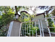 Eine zweistöckige Villa mit abgerundetem Dach hinter einem verwitterten weißen Metallzaun, umgeben von dichtem Grün und großen Bäumen; im Vordergrund ein Betonpfosten mit einer traditionellen schwarzen Laterne.