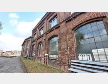 Backsteinfassade eines historischen Bahnhofsgebäudes mit großen, bogenförmigen Fenstern und Ziegelarchitektur. Im Vordergrund steht eine Holzbank, daneben verläuft ein gepflasterter Gehweg entlang der Schienen.