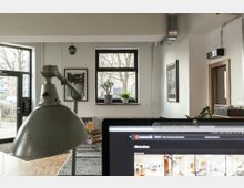 Moderner Büroraum mit großen Fenstern, die viel Tageslicht hereinlassen, einer Topfpflanze auf der Fensterbank und schwarzen gerahmten Bildern an der Wand. Im Vordergrund ist eine Schreibtischlampe und ein Laptop-Bildschirm sichtbar.