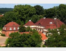 Historisches Gebäudeensemble mit roten Ziegeldächern, hellen Fassaden und barocken Details, umgeben von dichtem Grün und hohen Bäumen in einer ländlichen Umgebung.