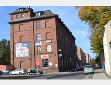 Backsteingebäude eines ehemaligen Industriebaus mit mehreren Etagen und großen Fenstern, heute mit verschiedenen Schildern und Werbeplakaten für Freizeitaktivitäten versehen. Das Gebäude steht an einer ruhigen Straße mit parkenden Autos und angrenzenden Wohnhäusern, umgeben von Bäumen.