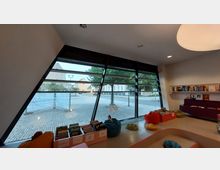 Innenansicht eines modernen Lesebereichs mit großen, schrägen Fenstern, die den Blick nach draußen auf einen gepflasterten Platz und umliegende Gebäude freigeben. Im Raum befinden sich bunte Sitzkissen, Bücherregale an der Wand und eine Auslage mit Kinderbüchern.