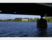 Blick von unter einer Brücke auf einen See, im Hintergrund ein großes, mehrstöckiges Gebäude mit blassem Putz und dunklem Dach, umgeben von Bäumen und Grünflächen.