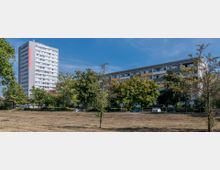 Ein moderner Wohnkomplex mit einem hohen Mehrfamilienhaus links und einem länglichen Gebäude rechts. Im Vordergrund befinden sich eine Rasenfläche und mehrere Bäume, die die Gebäude teilweise verdecken.