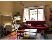 Kinderzimmer mit Etagenbett, rotem Sofa und hellem Holztisch in der Mitte. Große Fenster mit rosa Vorhängen lassen Tageslicht herein, und der Boden ist mit einem bunten Teppich bedeckt.