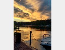 Sonnenuntergang an einem See mit ruhigem Wasser, reflektierendem Abendhimmel in warmen Gelb- und Orangetönen. Im Vordergrund ein Steg aus Backsteinen mit einem vertäuten Boot, im Hintergrund eine bewaldete Uferlandschaft.