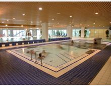 Hallenbad mit mehreren Becken, darunter ein Whirlpool im Vordergrund mit blauen Fliesen, und einem großen Schwimmbecken im Hintergrund. Der Raum hat eine Holzdecke, große Fenster und vereinzelte Pflanzen als Dekoration.