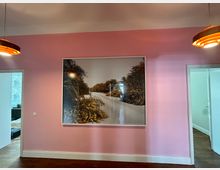 Ein Innenraum mit rosa gestrichener Wand, an der ein großes gerahmtes Landschaftsfoto hängt, das Büsche und einen schmalen Weg zeigt. Links und rechts der Wand sind Türöffnungen, die jeweils in angrenzende Räume mit hellen Wänden führen.