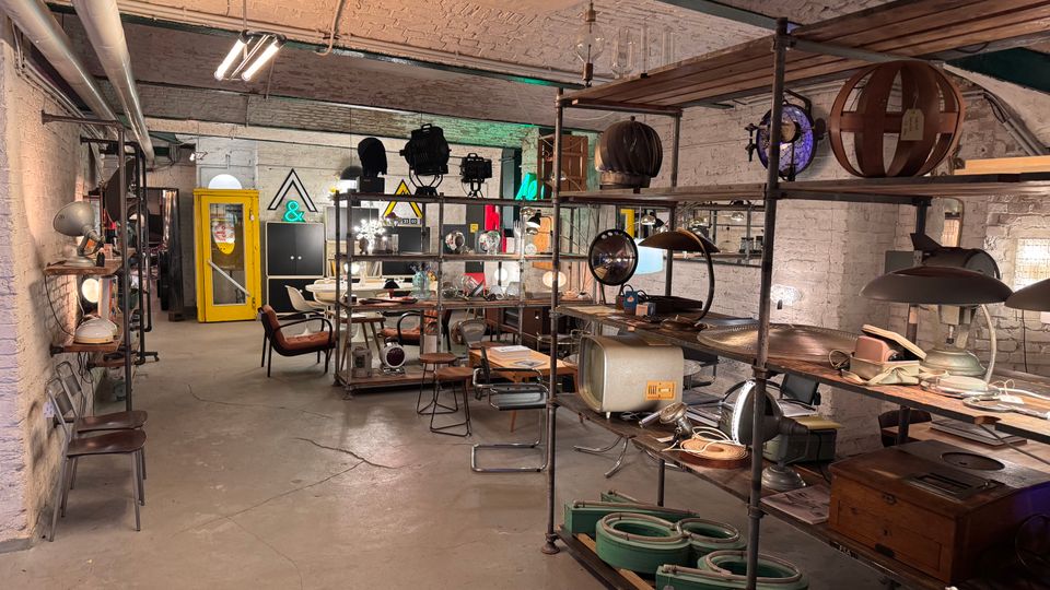 Industrie-ähnlicher Innenraum mit gewölbter Backstein-Decke, Regalen voller Vintage-Gegenstände wie Lampen, Elektrogeräte und Schilder; im Hintergrund eine gelbe Telefonzelle und dekorative Beleuchtung. Die Wände bestehen aus weiß gestrichenem Mauerwerk, und der Boden ist aus Beton.