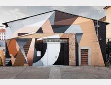 Industriegebäude mit einer Fassade aus Ziegelstein, kunstvoll bemalt in geometrischen Mustern mit braunen, schwarzen, weißen und grauen Farbtönen. In der Mitte befindet sich eine weiße Tür, daneben ein kleines Fenster mit Glasbausteinen und rechts eine Metallleiter, die an der Wand montiert ist.