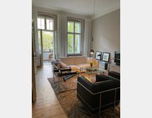 Wohnzimmer mit hellem Holzboden, einem weißen Sofa, einem schwarzen Ledersessel und einem gläsernen Couchtisch. Im Hintergrund sind große Fenster mit weißen Vorhängen sowie ein Zugang zu einem Balkon mit Blick auf grüne Bäume sichtbar.