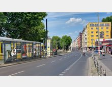 Straßenszene in einer Stadt mit Straßenbahnhaltestelle links, Bushaltestellenschild und gelber Straßenbahn. Rechts sind bunte Mehrfamilienhäuser und Geschäfte zu sehen, darunter ein Sparkassengebäude und Imbissstände. Eine breite Straße mit Fahrradweg führt durch die Szene, auf der Radfahrer und Fußgänger unterwegs sind.