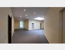 Leerer Innenraum eines Bürogebäudes mit hellbeigen Wänden, eingelassenen Deckenstrahlern und einer abgehängten Decke. Im Hintergrund befindet sich eine Glastür, die Tageslicht hereinlässt, sowie mehrere offene Türrahmen entlang der Wände.