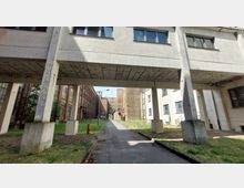 Industriegelände mit Gebäuden im Stil der frühen bis mittleren 20. Jahrhunderts. Links ein Backsteingebäude mit großen Fenstern, rechts ein hell verputztes Gebäude; ein betonierter Übergang verbindet beide Seiten, darunter ein schmaler Weg und grüne Rasenflächen.