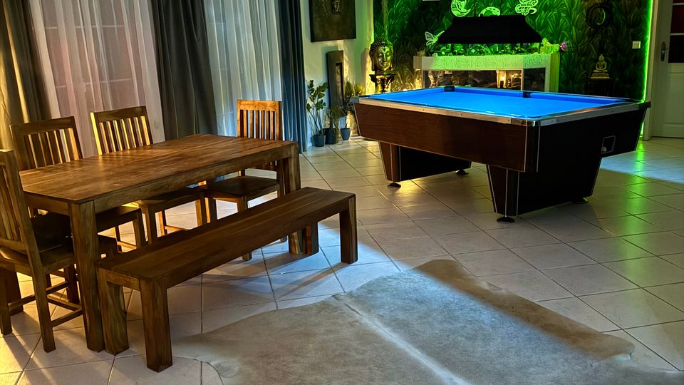 Großer Wohn- und Spielbereich mit gefliestem Boden, Esstisch aus Holz mit Bänken und Stühlen sowie einem Billardtisch. Im Hintergrund hängen bodentiefe Vorhänge; eine grün beleuchtete Wand mit Dschungelmotiv und Kamin bildet einen auffälligen Akzent.