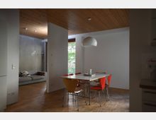 Wohnraum mit Holzboden und deckenhoher Trennung in zwei Bereiche; ein Essbereich mit einem rechteckigen Tisch, umgeben von gelben und roten Stühlen, sowie eine Hängelampe darüber, und ein Sofa im Hintergrund neben einer Betonwand und einem großen Fenster mit Blick ins Grüne.