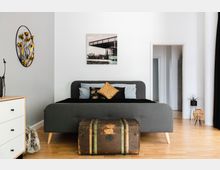 Schlafzimmer mit einem grauen Polsterbett, dekoriert mit Kissen in verschiedenen Mustern und Farben. Vor dem Bett steht eine antike Holztruhe, an der Wand hängt ein Metall-Wanddekor mit Blütenmotiv, und ein heller Holzboden ergänzt das moderne und minimalistische Design.