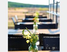 Außenbereich eines Cafés oder Restaurants mit Holztischen und schwarzen Stühlen, auf denen kleine Glasvasen mit frischen Blumen stehen; im Hintergrund ist eine ländliche Landschaft mit Bäumen und Wiesen zu sehen.