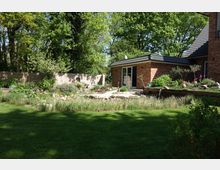 Ziegelhaus mit einem modern gestalteten Garten, der aus einem gestuften Steingarten, dicht bepflanzten Beeten und einer Rasenfläche besteht. Im Hintergrund sind eine Holz- und eine Ziegelmauer sowie Bäume zu sehen, die den Garten umgeben.