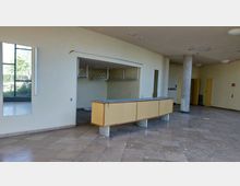 Innenraum eines verlassenen Gebäudes mit einem leeren Empfangstresen aus Holz in der Mitte, einer Garderobenstange im Hintergrund und abgenutztem Boden aus Fliesen. An der Decke sind beschädigte Stellen sichtbar, daneben befinden sich große Fenster und mehrere Türen.