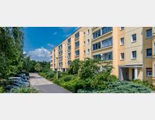 Mehrstöckiges Wohngebäude mit gelber Fassade und großen Fenstern, umgeben von gepflegten Grünanlagen und geparkten Autos entlang einer gepflasterten Zufahrtsstraße unter blauem Himmel mit wenigen Wolken.