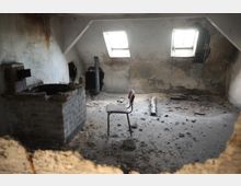 Ein verlassenes und stark beschädigtes Zimmer mit einer Beton- und Ziegelstruktur. Die Wände sind schmutzig und teilweise eingerissen, zwei kleine Dachfenster lassen Licht herein, und in der Mitte steht ein einzelner Stuhl auf einem Boden voller Schutt.