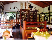 Gemütliches Restaurant im Landhausstil mit Holzmöbeln, gedeckten Tischen und rustikaler Dekoration an den Wänden, wie antiken Werkzeugen, Jagdtrophäen und Wandmalereien. Helle Wände und eine hölzerne Trennwand unterteilen den Raum, der von natürlichem Licht und Hängeleuchten erhellt wird.