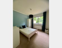 Helles Schlafzimmer mit weißem Bettgestell, Kleiderschrank und Kommode, einer blau gestrichenen Akzentwand und Teppichboden. Großes Fenster mit dunklen Vorhängen und Heizkörper darunter, Blick ins Grüne.