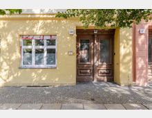 Fassade eines Wohnhauses mit gelber Wand, einem weiß gerahmten Fenster links und einer hölzernen Doppeltür rechts. Der Gehweg davor ist mit Kopfsteinpflaster gestaltet, umgeben von Schatten von Bäumen.