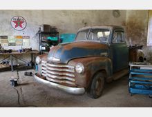 Ein alter, rostiger Pickup-Truck steht in einer Werkstatt mit bröckelnden Wänden. Im Hintergrund sind ein Werkbank mit Werkzeugen, ein Texaco-Werbeschild und an der Wand befestigte Kennzeichen zu sehen.