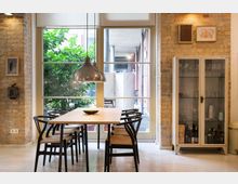 Interior dining area with exposed brick walls, a wooden table and black chairs beneath a hanging metal pendant light. Large floor-to-ceiling glass doors and windows open to a small courtyard with greenery, and a white glass-front cabinet displays glassware and bottles.