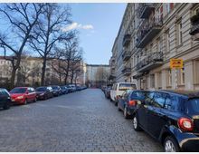 Straßenszene in einem Wohngebiet mit Kopfsteinpflaster, parkenden Autos auf beiden Seiten und mehrstöckigen Altbauhäusern mit Balkonen. Links stehen kahle Bäume, und im Hintergrund sind weitere Gebäude sichtbar.