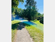 Ein geschwungener Schotterweg führt durch eine sonnige Rasenfläche mit hohen Bäumen. Im Hintergrund verläuft eine steinerne Balustrade mit dekorativen Elementen entlang des Parks.