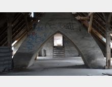 Ein verlassener Dachboden mit sichtbaren Holzbalken und einem großen betonierten Spitzbogen in der Mitte. Die Betonwand und andere Oberflächen sind mit Graffiti bedeckt, und eine kleine Holztreppe führt zu einem Lichtdurchlass im Hintergrund.