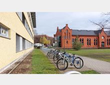 Gelände mit einem modernen, gelbbraunen Gebäude links und einem historischen, roten Backsteingebäude mit großen Rundbogenfenstern rechts; im Vordergrund mehrere geparkte Fahrräder an einem Fahrradständer und eine gepflasterte Fläche, umgeben von Grünflächen.