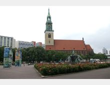 Eine historische Kirche mit einer markanten grünen Turmspitze und einem roten Ziegeldach steht im Hintergrund. Im Vordergrund befinden sich der Neptunbrunnen mit Skulpturen und Wasserspielen sowie Blumenbeete mit roten Rosen, umgeben von Bäumen und modernen Gebäuden.