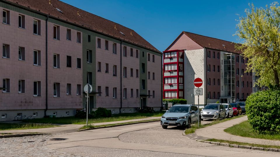 Straßenszene in einem Wohngebiet mit mehrstöckigen, verputzten Mehrfamilienhäusern und roten Fensterrahmen sowie einem gläsernen Treppenhausanbau. Entlang der kurvigen Fahrbahn stehen geparkte Autos, daneben Gehwege, Grünflächen und ein Straßenschild „Joliot-Curie-Straße“.