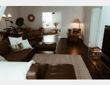 Wohnzimmer mit dunkelbraunem Holzboden, einer braunen Ledercouch und einem beigefarbenen Teppich; im Hintergrund ein Essbereich mit Holztisch und Stühlen sowie ein angrenzender Schlafbereich mit weißer Bettwäsche und dekorativen Pflanzen.