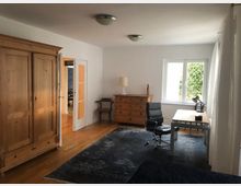 Helles Zimmer mit Holzboden, ausgestattet mit einem großen Holzkleiderschrank, einer Holzkommode, einem Schreibtisch mit Drehstuhl und einem Fenster, das auf Grünblick zeigt. Im Hintergrund ist eine Tür mit Glaseinsatz sichtbar, die zu einem anderen Raum führt.