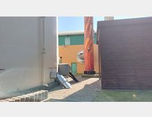 Industrieanlage mit mehreren Gebäuden und Anlagenkomponenten. Im Vordergrund sind weiße und braune Strukturen sowie ein dekorativ bemalter Schornstein zu sehen; im Hintergrund eine gelb-orange Backsteinfassade mit grünem Tor.