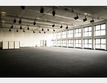 Großer, leerer Veranstaltungssaal mit dunklem Teppichboden, hohen weißen Wänden und großen, bodentiefen Fenstern, die viel Tageslicht hereinlassen. An der Decke sind mehrere Scheinwerfer und Lichtstreben angebracht.