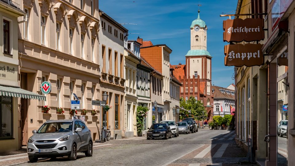 Blick entlang einer schmalen Altstadtstraße mit Kopfsteinpflaster, historischen Fassaden und kleinen Geschäften mit Ausleger-Schildern. Am Straßenende ragt ein hoher Kirchturm mit grüner Kuppel über die Dächer.